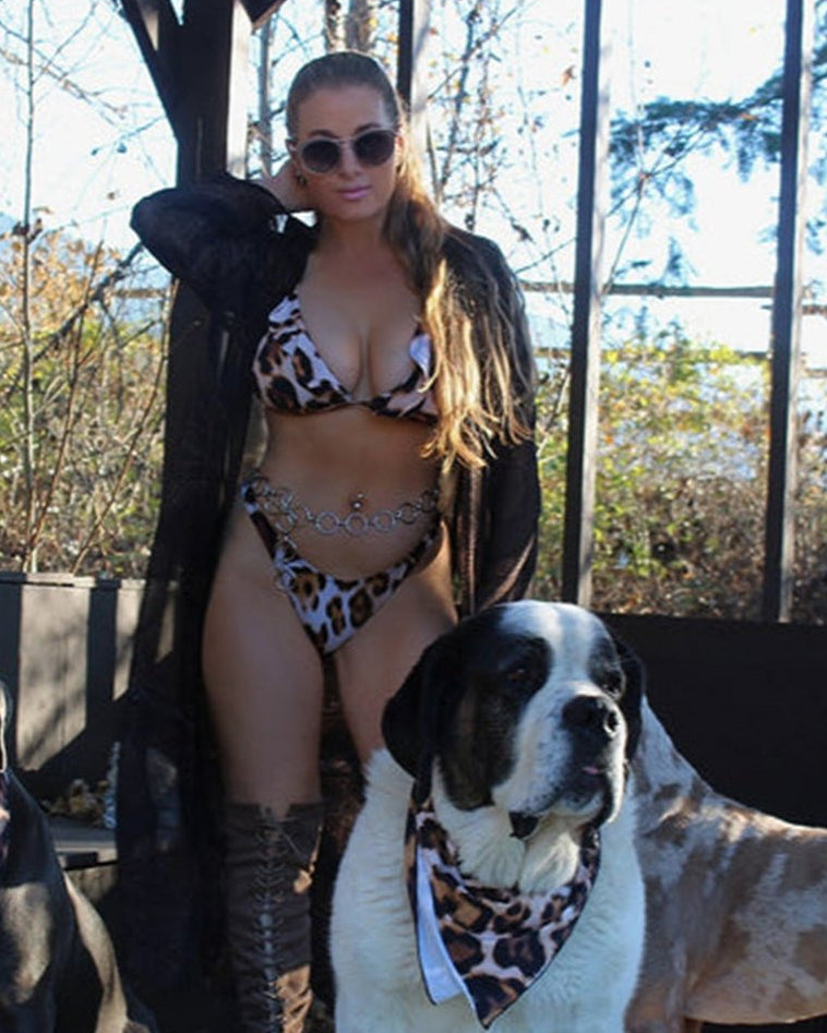 A woman wearing a leopard print bikini with a triangle top and cheeky bottoms, standing next to two dogs wearing matching bandanas.