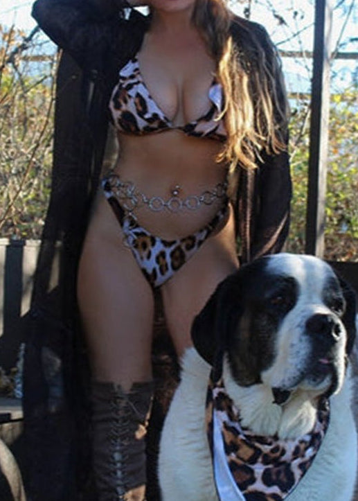 A woman wearing a leopard print bikini with a triangle top and cheeky bottoms, standing next to two dogs wearing matching bandanas - Jaiskini swimwear