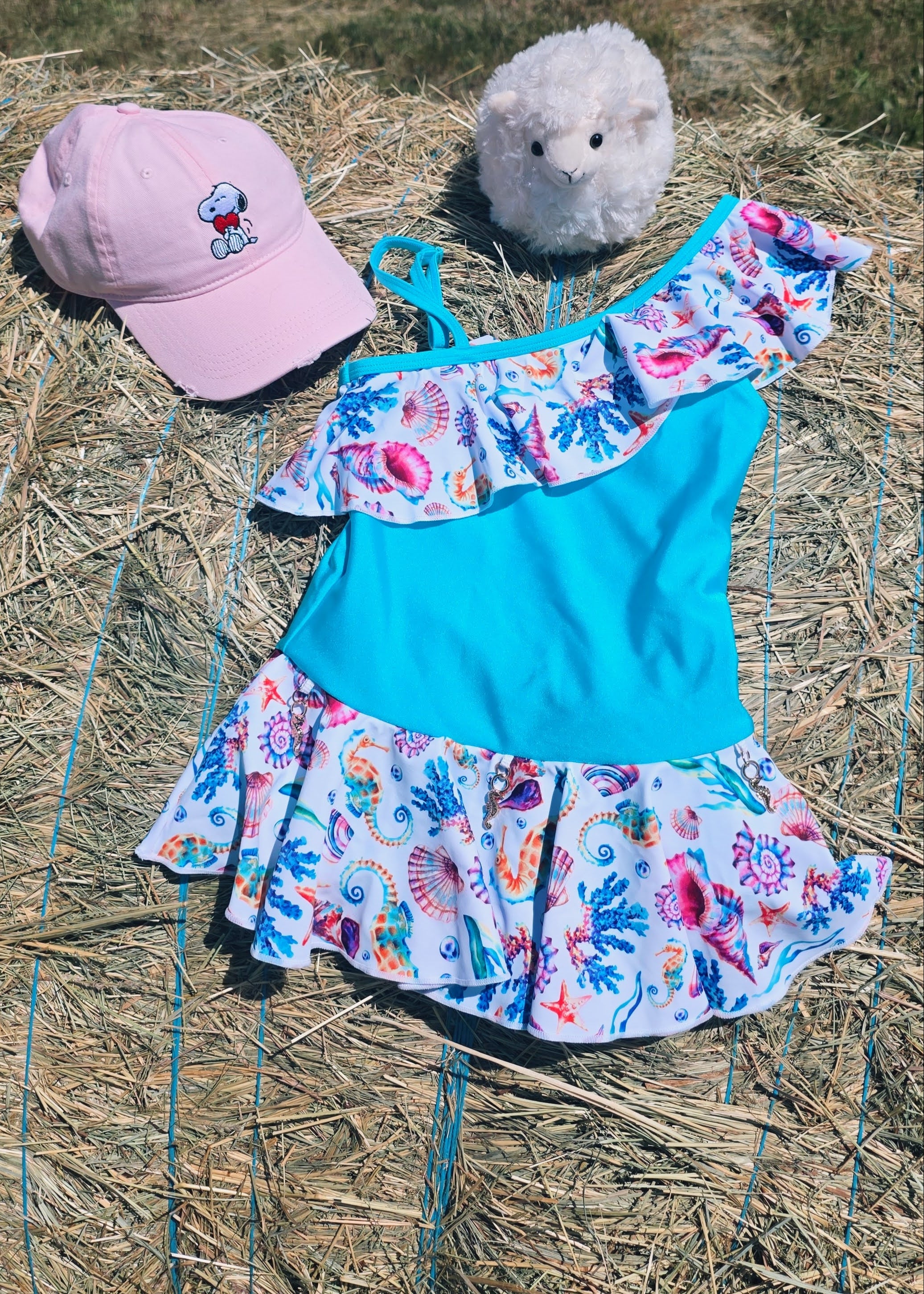 Children's floral dress with blue top and colorful skirt on a straw background, accompanied by a pink cap and plush toy.